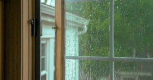 Rain droplets on a window with green trees and neighboring buildings visible outside.